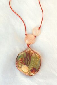 Pendentif résine forme organique sur cordon de soie rouge avec inclusion de feuille eucalyptus, coquillages, strass, pétale de rose, gorgone rouge, personnage en fil laiton, le tout représentant un paysage de songe